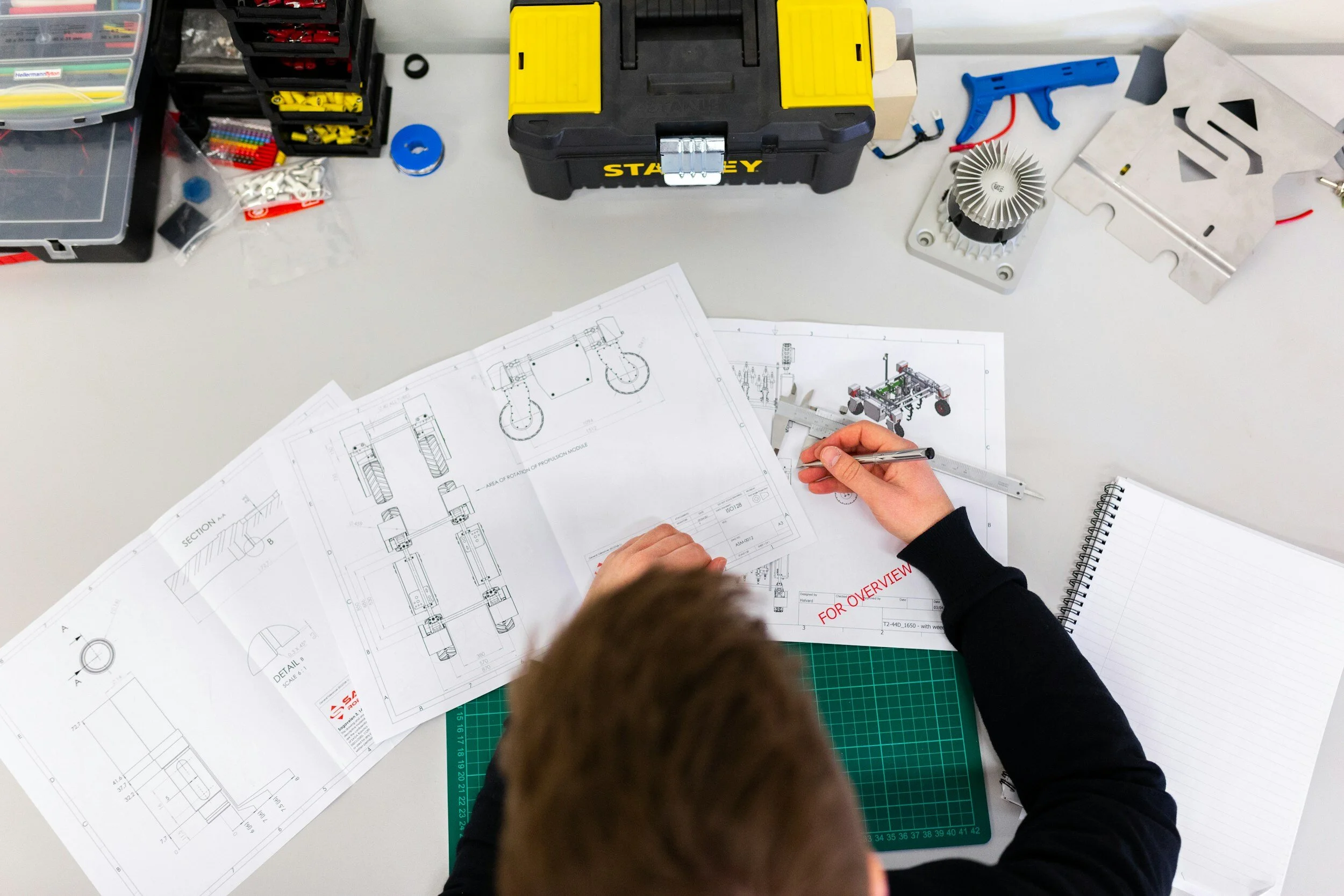 Engineer measuring a mechanical part with calipers over printed engineering blueprints on a workbench